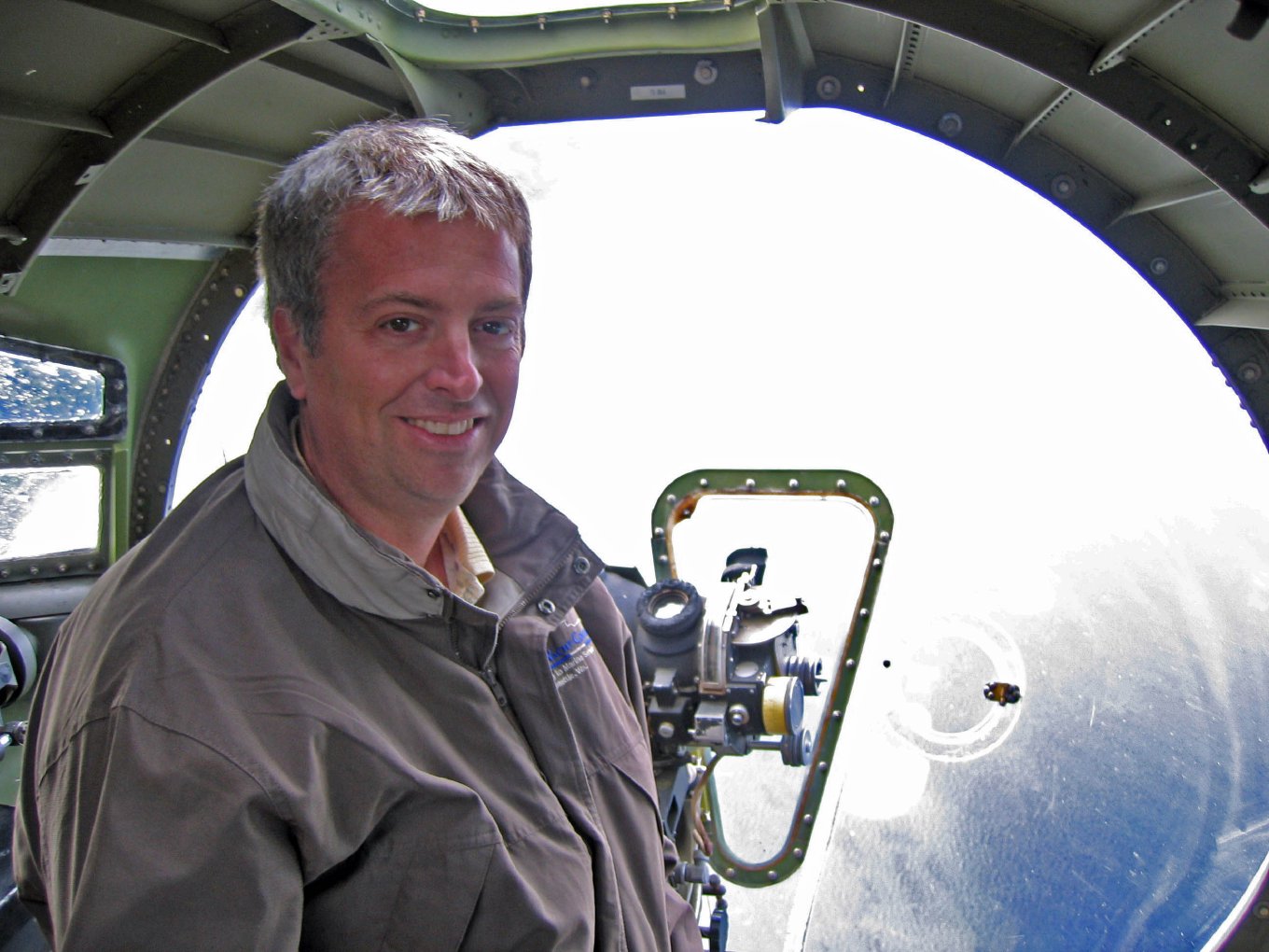 Brian smiling beside the Norden bombsight in the plexiglass nose