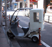 A three-wheeled scooter with a roof and a cargo box, parked on a sidewalk