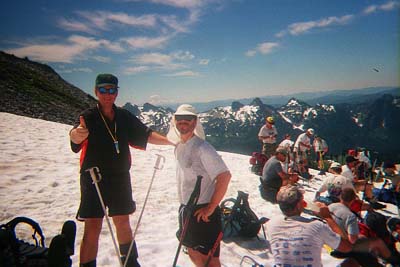 Mike and Jim in shorts and T-shirts, standing on the snow with the rest of the group resting behind them