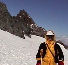 Mike in a yellow parka on the snow, with dark rock ridges behind him