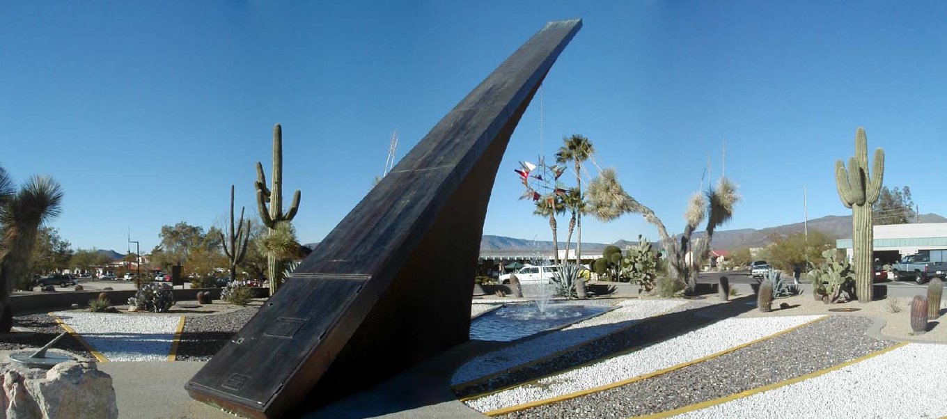 A panorama of a huge curved sundial blade rising from a traffic circle among cactus