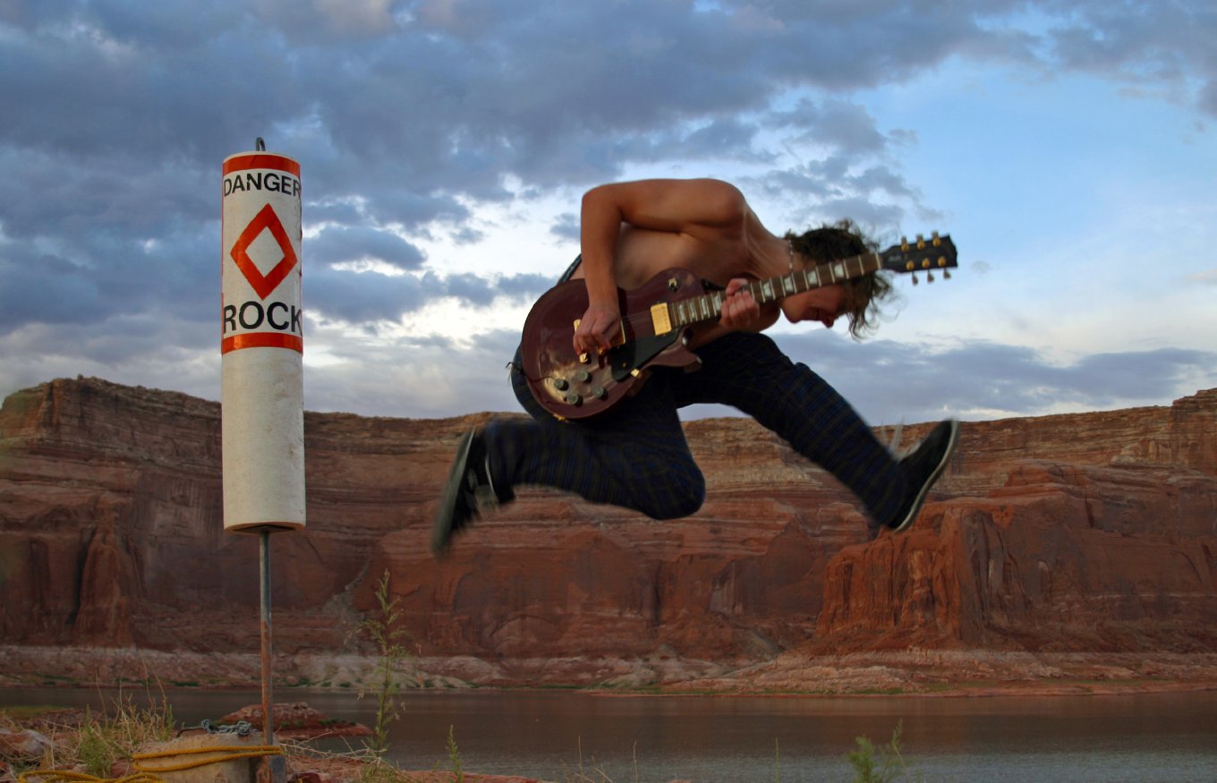 Chris leaping in the air with a guitar beside a Danger Rock marker, red canyon walls behind