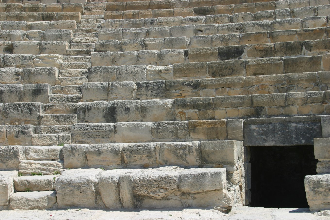 Tiers of worn limestone seats in an ancient amphitheater, with a dark doorway