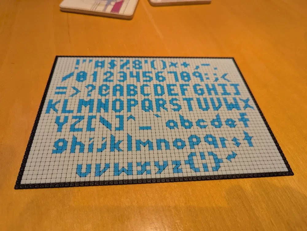 A 3D-printed tile sign showing the full character set in blue tiles on white, with a black border